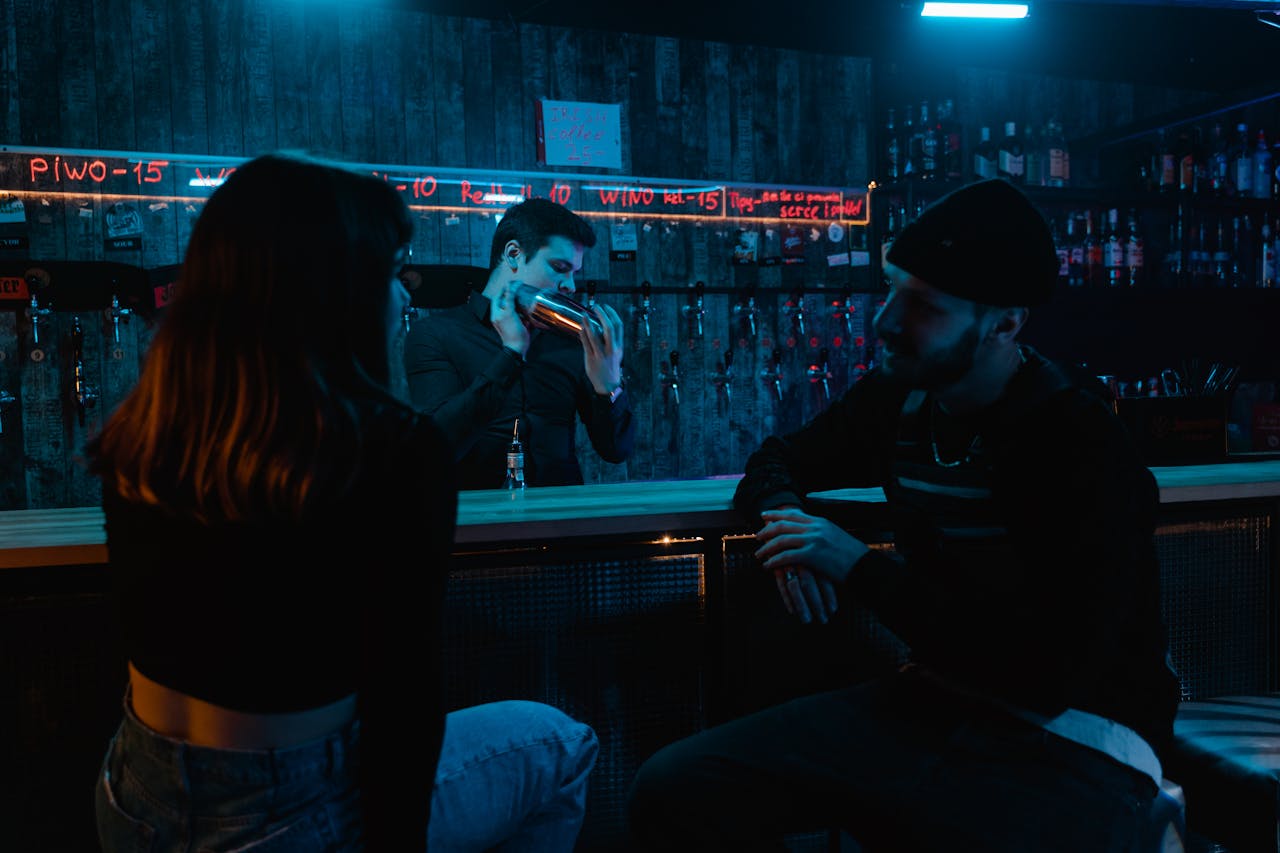 Intimate bar scene with people socializing and bartender preparing drinks under neon lights.